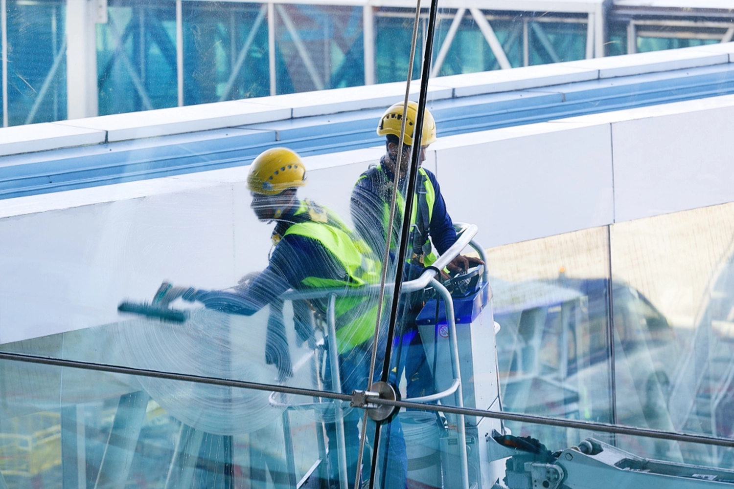 Duka Reinigung - Gebäudereinigung - Zwei Fensterputzer mit gelben Schutzhelmen und Warnwesten reinigen die Glasfassade eines Gebäudes von einer Hubarbeitsbühne aus.