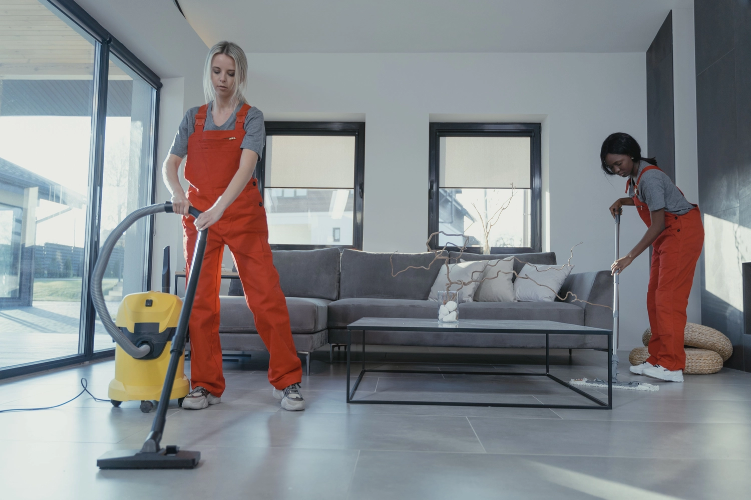 Duka Reinigung - Gebäudereinigung - Zwei Profis in Overalls saugen und reinigen ein modernes Wohnzimmer.