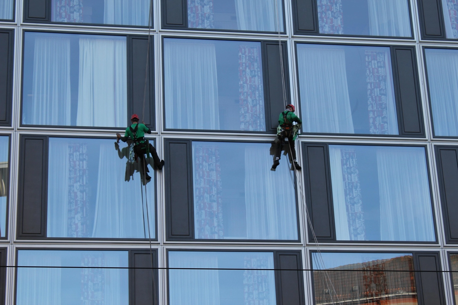 Duka Reinigung - Gebäudereinigung - Zwei Fensterputzer hängen an Seilen, um ein Hochhaus aus Glas zu reinigen.