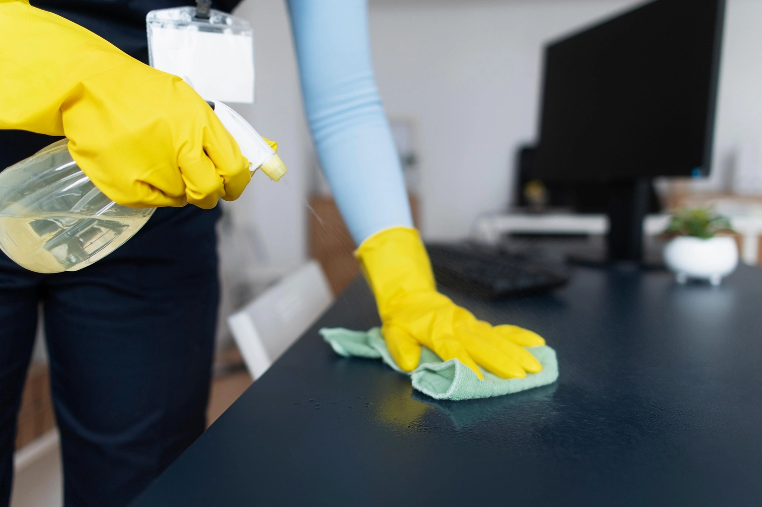 Duka Reinigung - Gebäudereinigung - Duka Reinigung - Gebäudereinigung - Eine Person mit gelben Handschuhen wischt einen Schreibtisch mit einem Tuch und einer Sprühflasche ab.